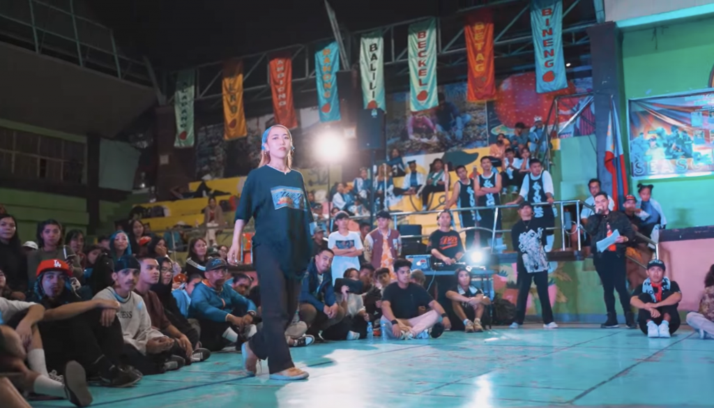 Nono Sasai, a young woman wearing a black shirt and headband walks confidently across a brightly lit stage, surrounded by an attentive crowd seated on the floor and in the bleachers. The venue is decorated with colorful banners displaying place names such as "Beckel" and "Balili." Spectators of various ages watch intently, and a man holding a microphone stands to the side, announcing the event. The atmosphere appears vibrant and energetic.